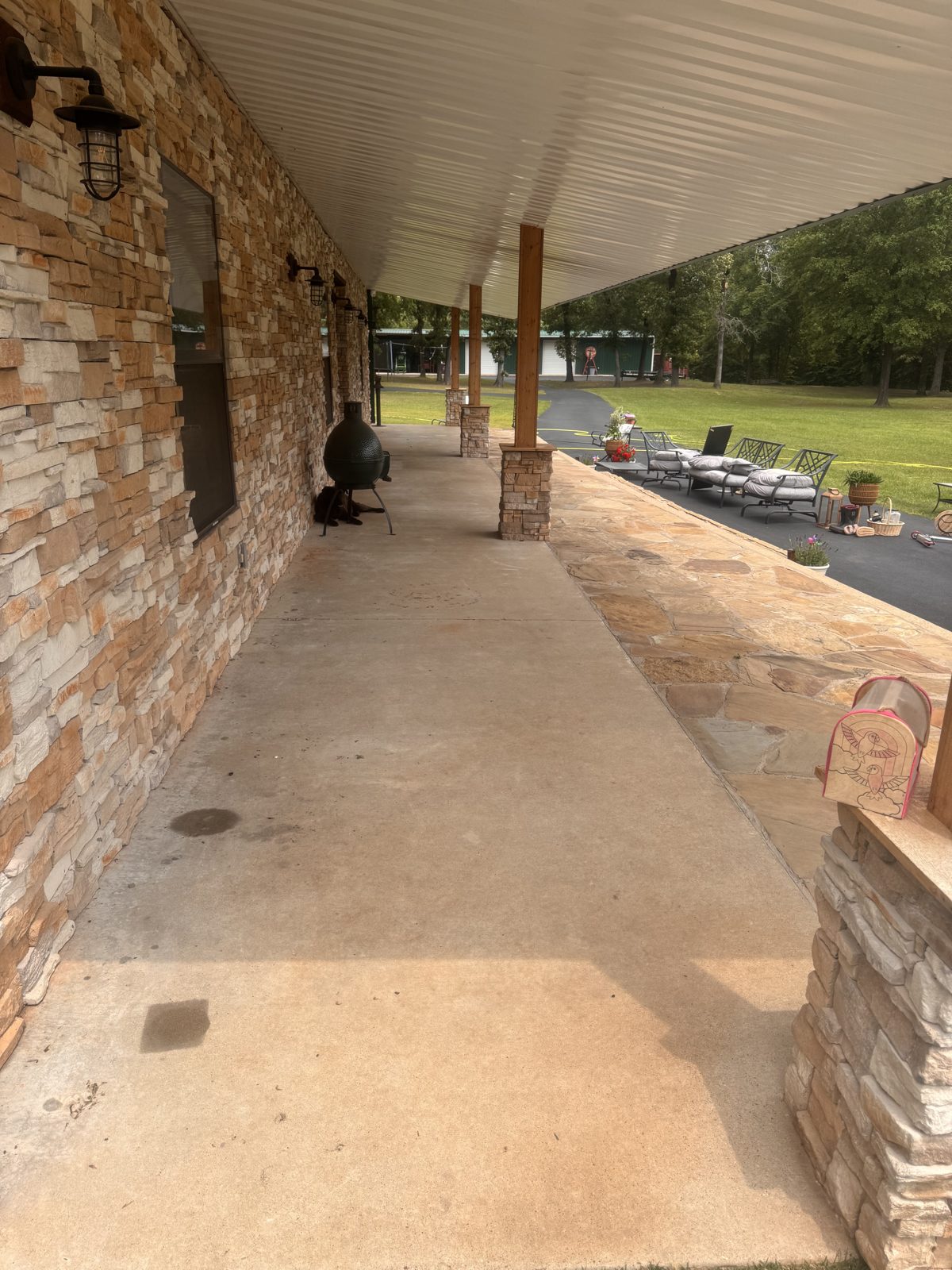 Long covered porch with stacked stone veneer wall and stone-wrapped posts, concrete porch floor with a few dark spots, flagstone walk alongside, wall lanterns, outdoor furniture on the drive