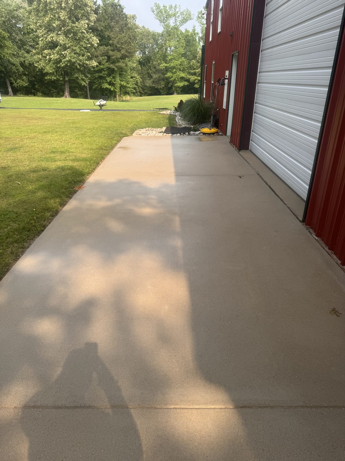 Long concrete slab beside a red metal shop, freshly washed and still wet, the photographer's shadow in the foreground, a yellow piece of equipment by the wall, lawn and trees at left